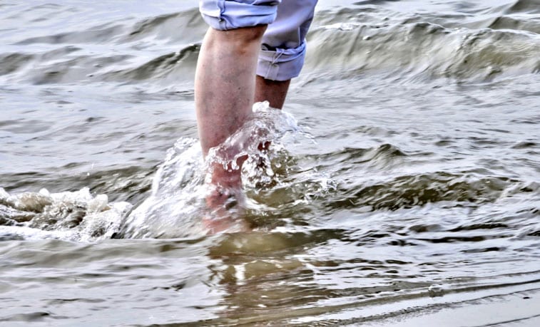 a man standing in the ocean, his jeans rolled up, revealing varicose veins