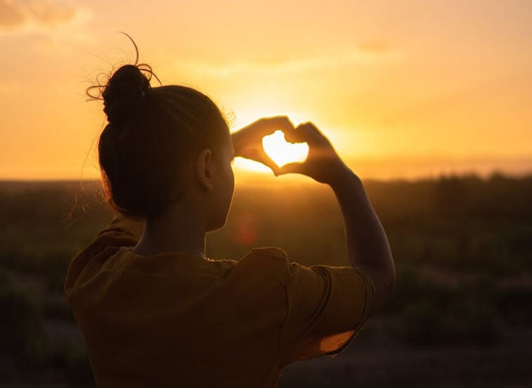 a woman holds her hand to form a heart, the sun shining through it